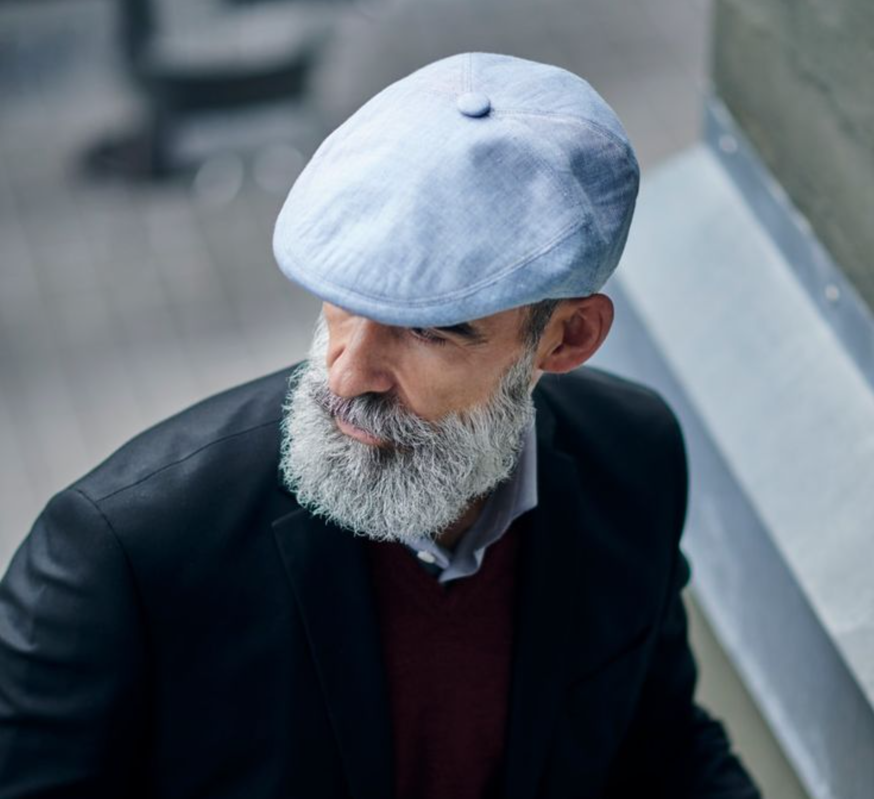 A stylish older man with a full gray beard wearing a light blue flat cap, a black blazer, and a burgundy sweater, looking down thoughtfully in an urban setting.
