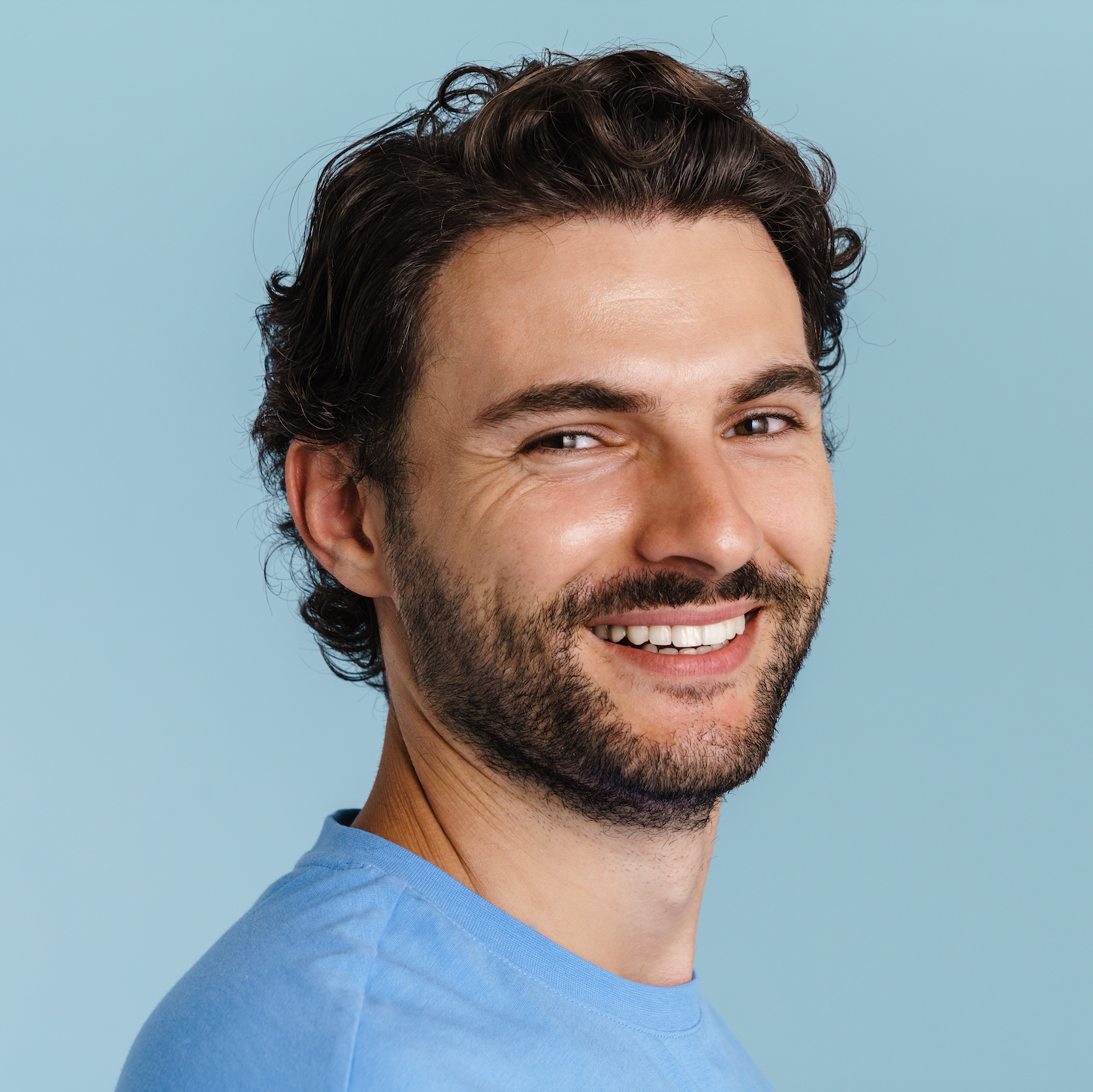 A smiling man with wavy dark hair and a beard, wearing a light blue t-shirt, looking over his shoulder against a pale blue background.
