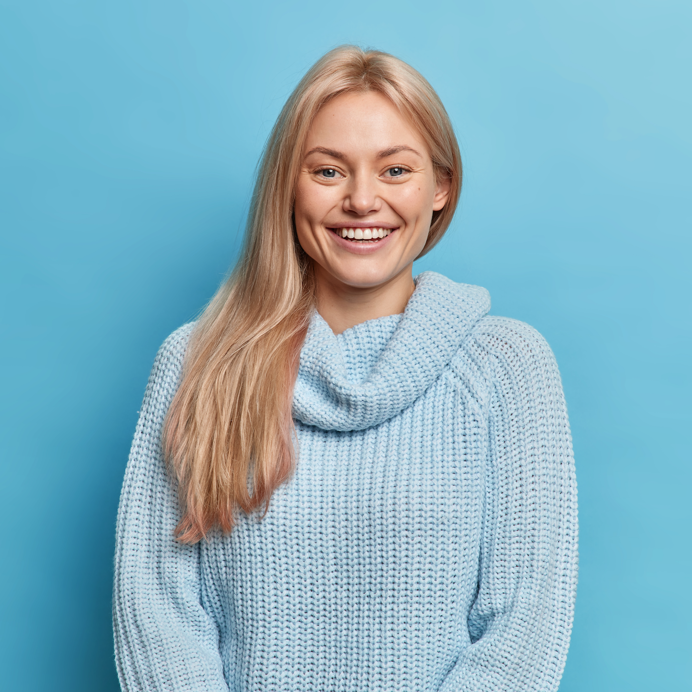 A cheerful blonde woman wearing a light blue knitted sweater, smiling brightly against a solid blue background.