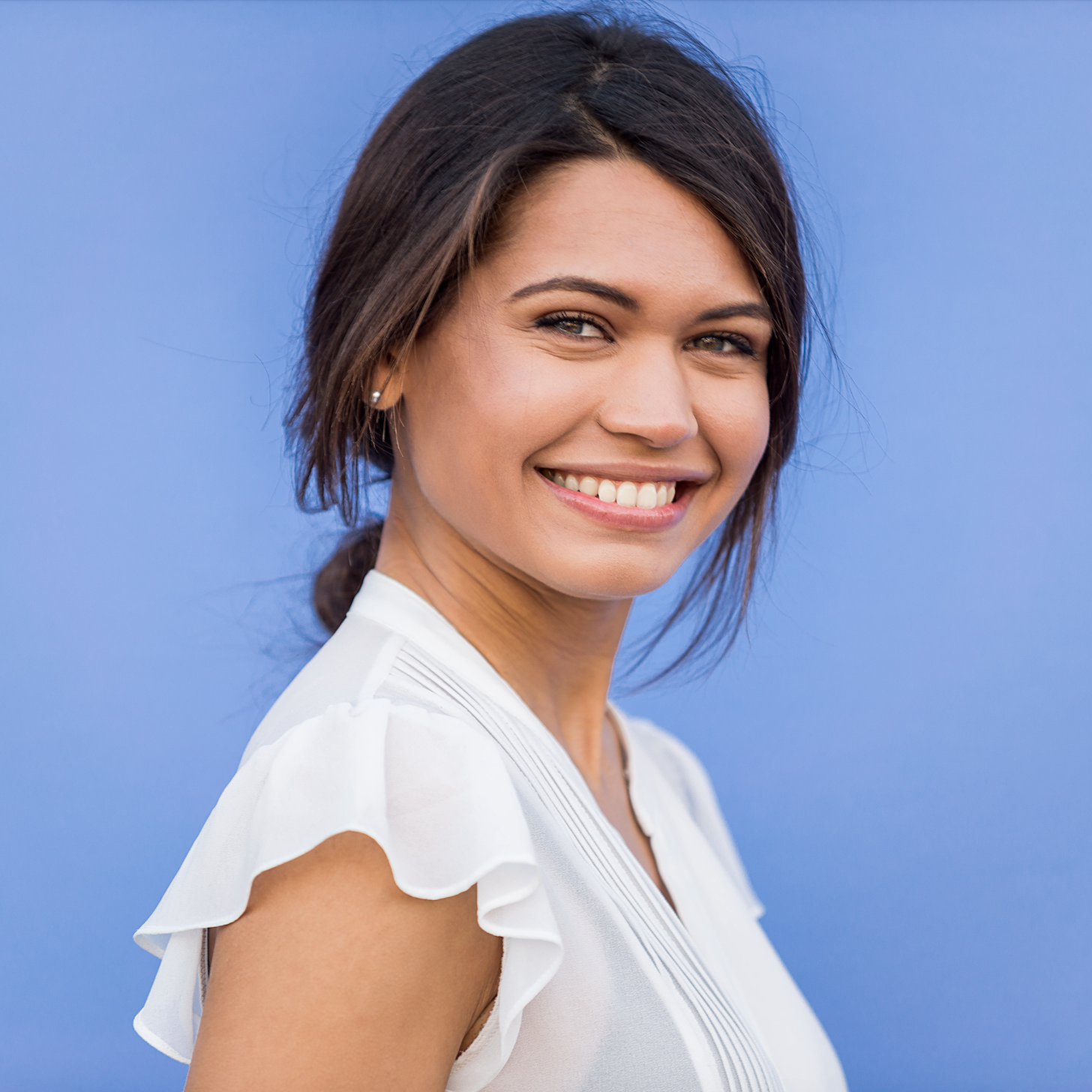 A smiling woman with dark hair tied back, wearing a white ruffled-sleeve blouse, posing against a solid medium blue background.