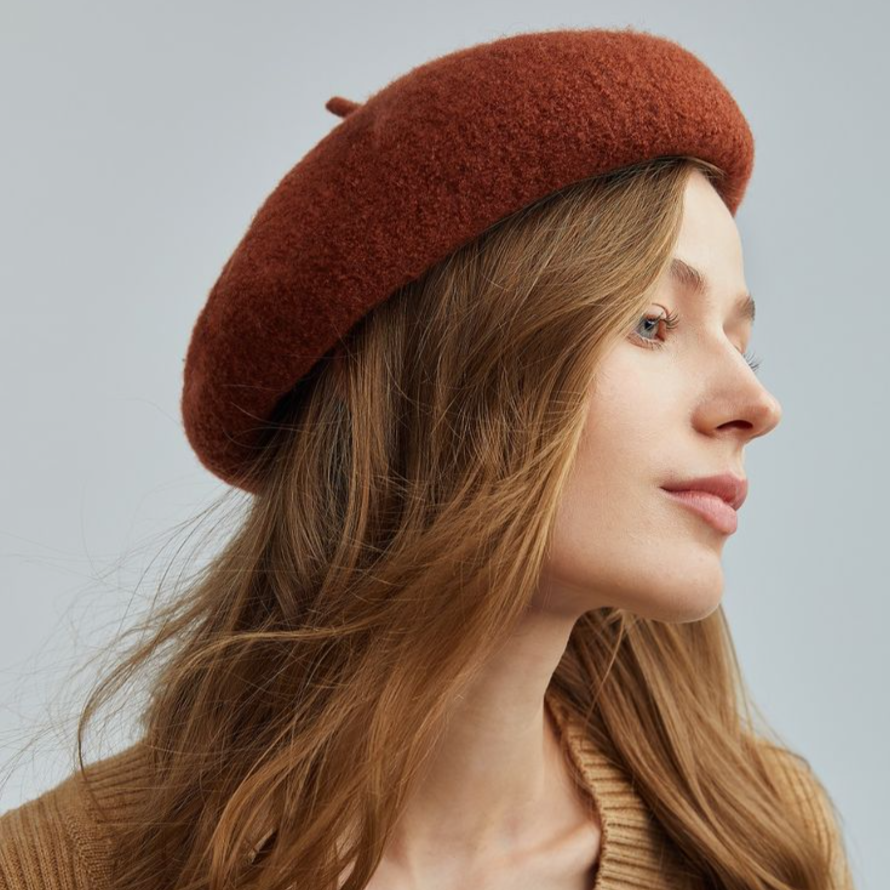 A woman with long light brown hair wearing a textured rust-colored wool beret, captured in a soft side profile against a light grey background.